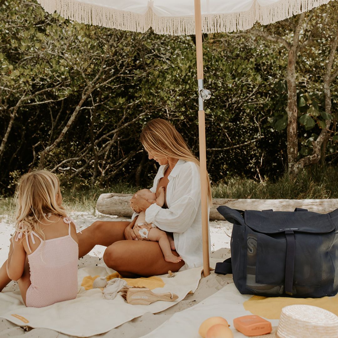 A mum sits on a towel under a beach umbrella, breastfeeding her newborn while chatting with her little girl. Next to them is the Sande Beach Hauler mesh backpack in Deep Sea Navy.