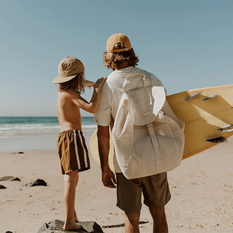 Dad and son checking the surf, with dad carrying a yellow surfboard and the Sande Beach Hauler mesh backpack, and son wearing a Sande merch cap.