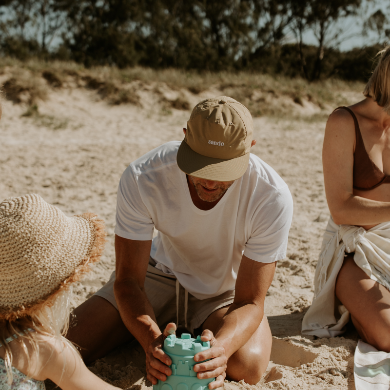 A lifestyle image of dad wearing the beige waterproof Sande cap while building sandcastles on the beach with his family.