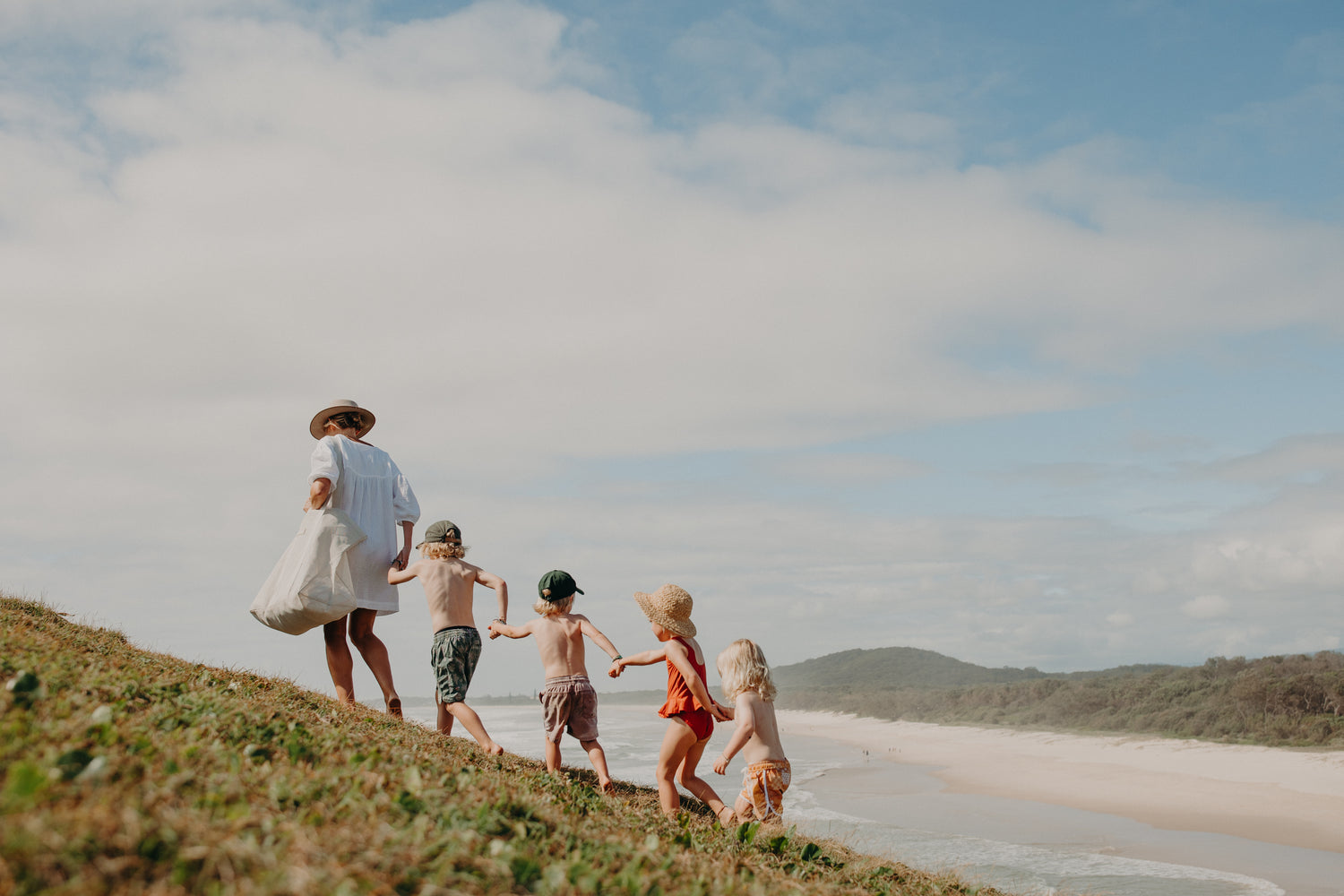 A mother with four children walking up a grassy headland with the beach and ocean in the background. Mum is carrying the oversized Sande Beach Hauler bag. Four children follow behind her. They are all holding hands.