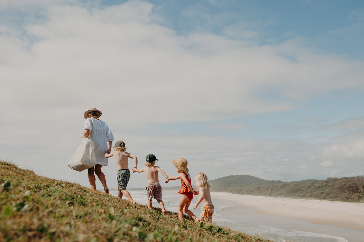 A mother with four children walking up a grassy headland with the beach and ocean in the background. Mum is carrying the oversized Sande Beach Hauler bag. Four children follow behind her. They are all holding hands.