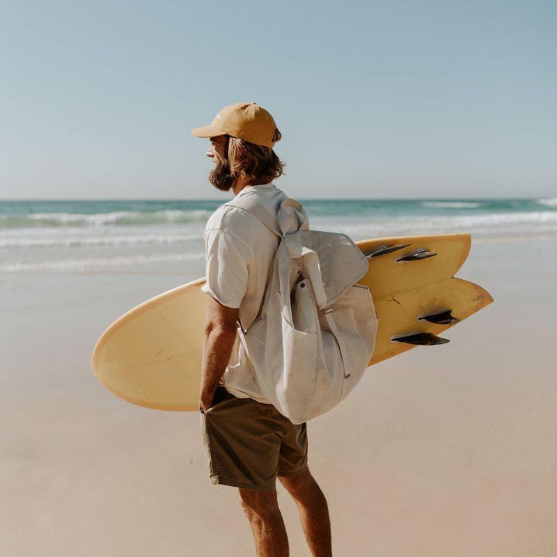Dad at the beach with Sande Beach Hauler mesh backpack on his back and yellow surfboard under one arm, ready for a beach day.