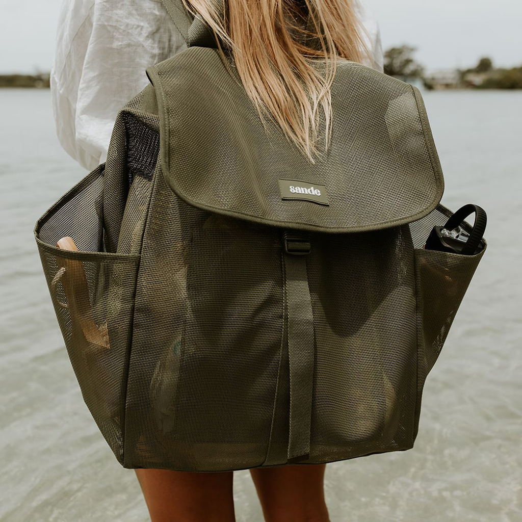 An image of a woman with long blonde hair standing at the water edge, wearing the Sande Beach Hauler mesh backpack in Pandanus, a dark olive green. The backpack features side pockets, a flap that clips up, and displays a silicone Sande logo in white.