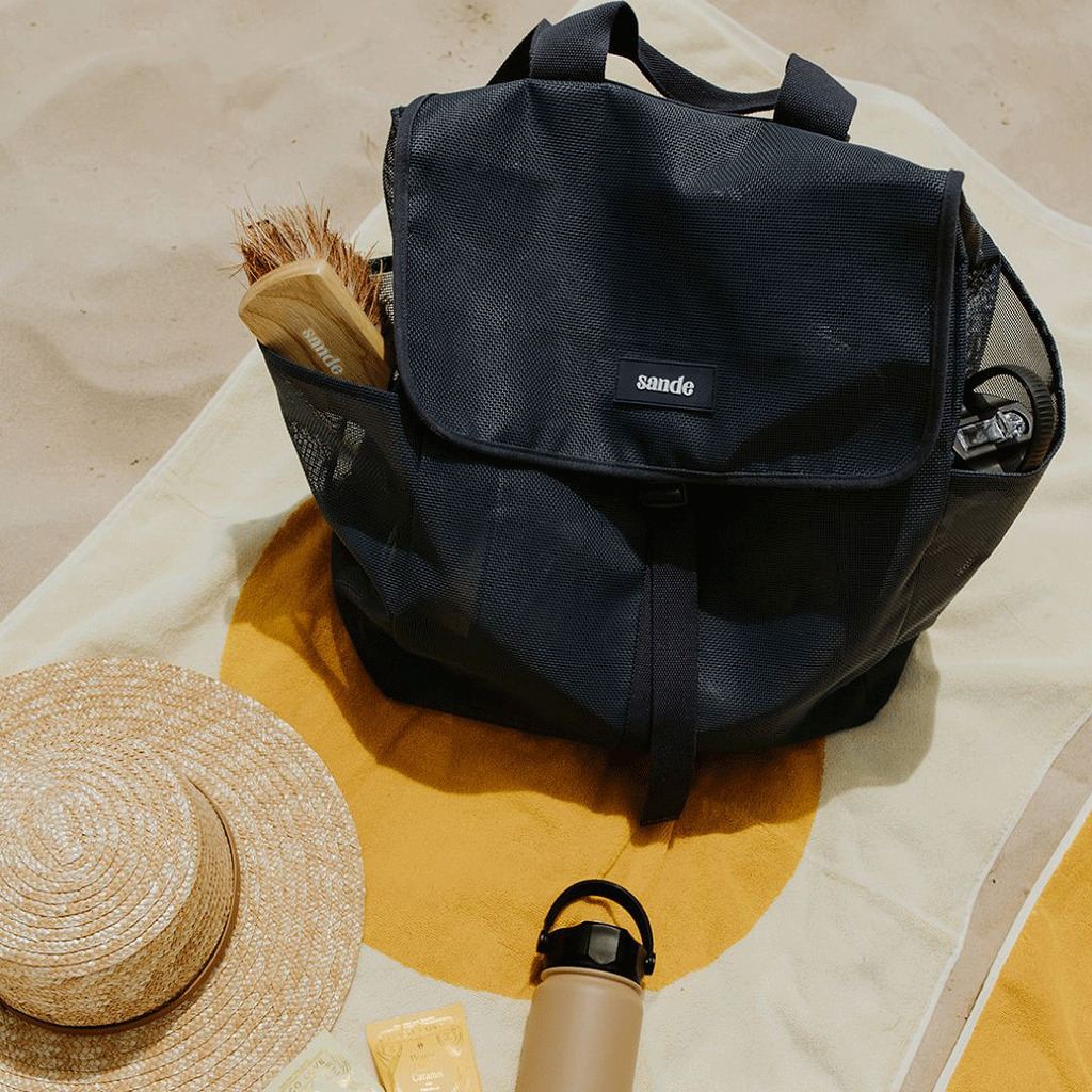 Top view of the Sande Beach Hauler mesh backpack, closed and sitting on a beach towel on the sand. A Pargo drink bottle and a Sande sand brush are placed in the outer mesh pockets.