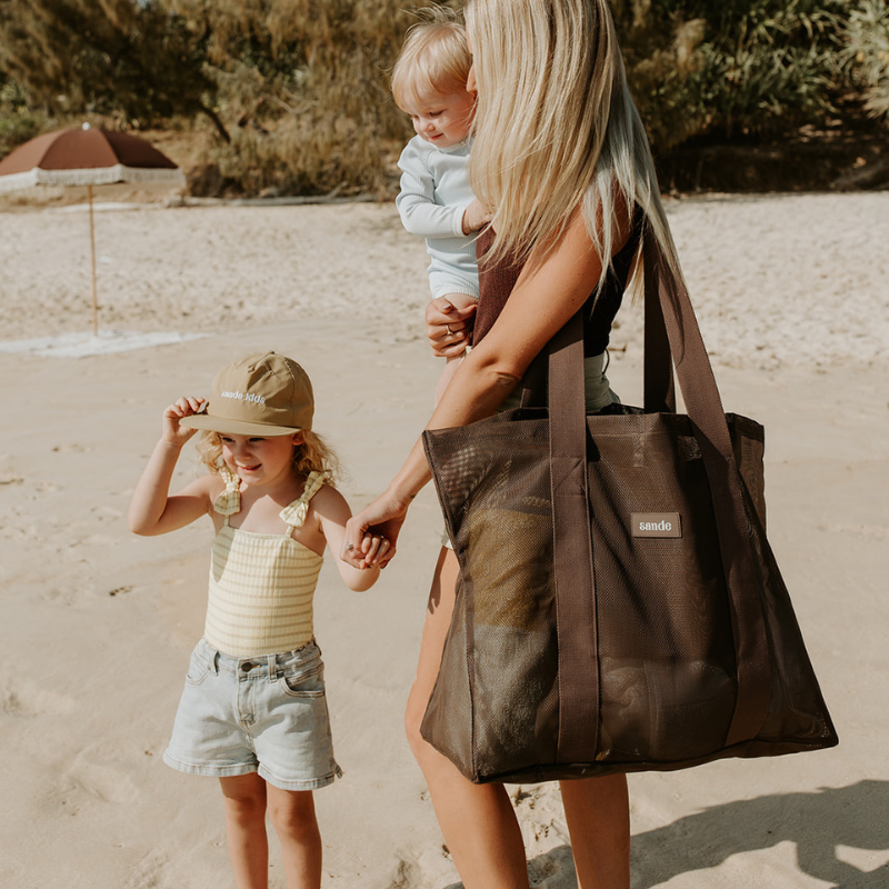 Woman holding a child and walking with a young girl on the beach, carrying the espresso brown Sande mesh beach bag tote.