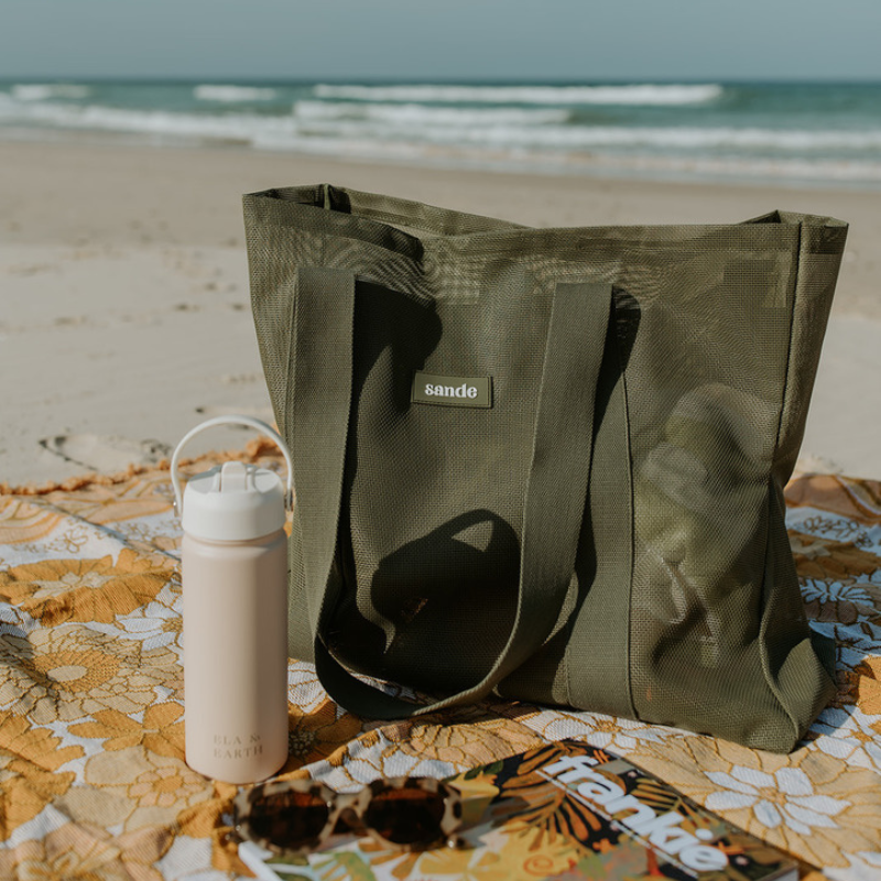 The Sande Beach Hauler - Mini mesh beach bag in Pandanus olive green, sitting on a yellow floral beach blanket at the beach. Ocean in the background.