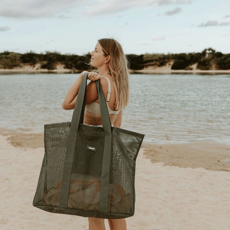 A women holding the Sande™ Beach Hauler oversized mesh beach bag in Pandanus green at a lake beach with dune sin the background.