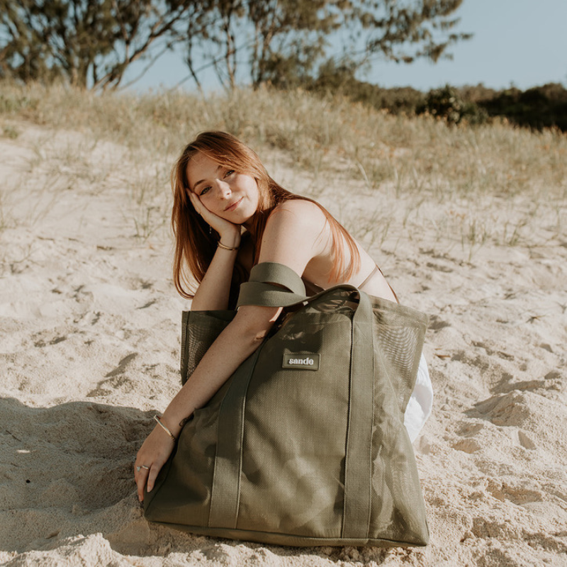 Young woman with long red hair squatting behind the Sande Beach Hauler Mini mesh beach bag in Pandanus Green at the beach, highlighting the spacious size of this large carry-all bag for beach essentials.