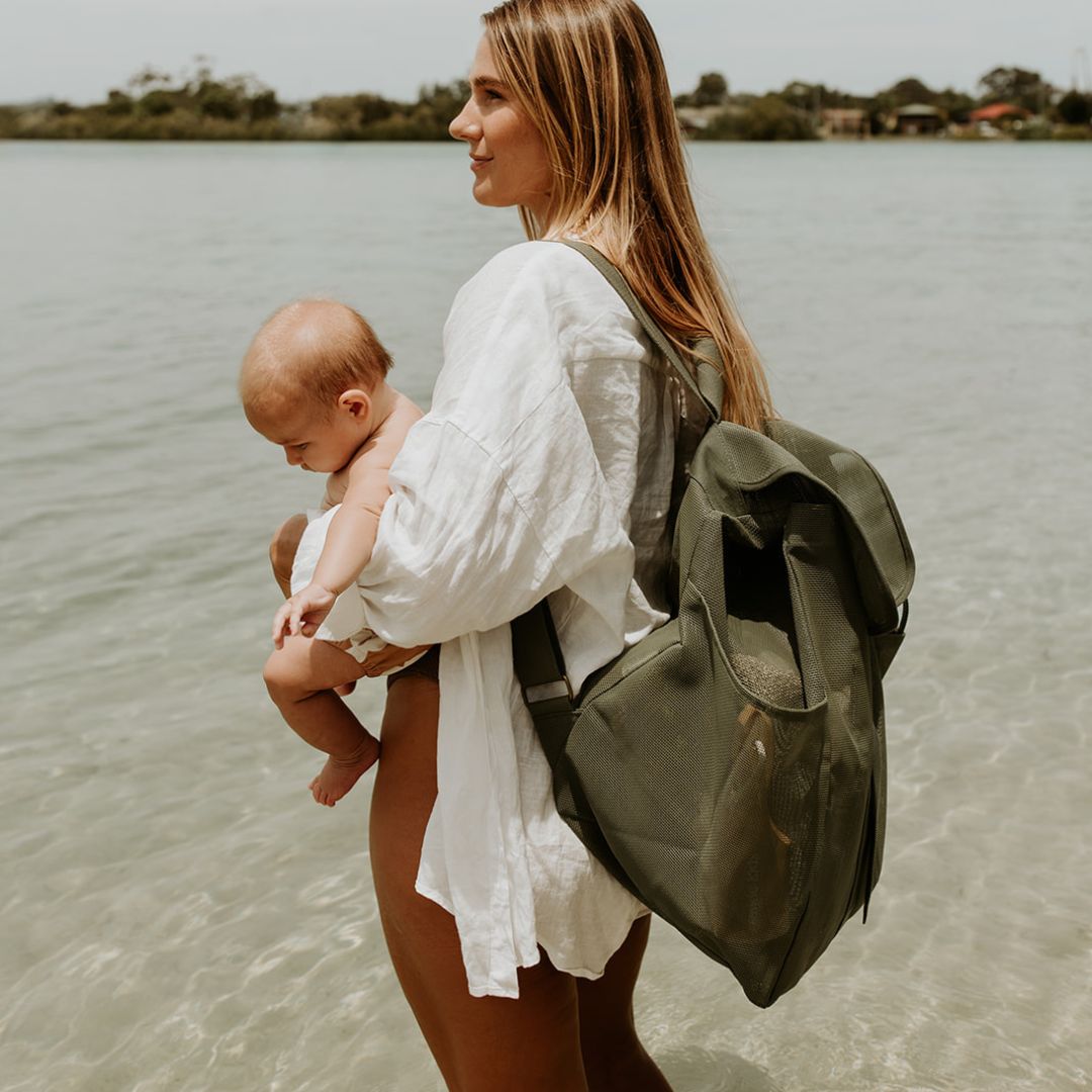 A mum with long blonde hair stands on the side at the waters edge, showcasing the side view of the dark olive green Sande Beach Hauler mesh backpack. She is holding a newborn baby in her arms. The backpack features side pockets, a flap with a clip, and a white silicone logo, making it both stylish and functional for family outings at the beach.