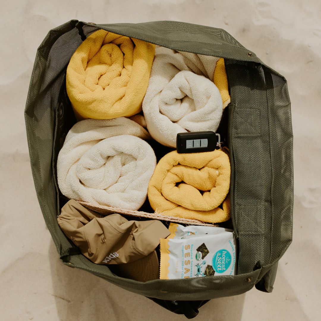 This image shows a top view of the Sande Beach Hauler mesh beach bag in dark green. The bag is packed with four bulky beach towels, hats, snacks, and other beach essentials, demonstrating its spacious and functional design.
