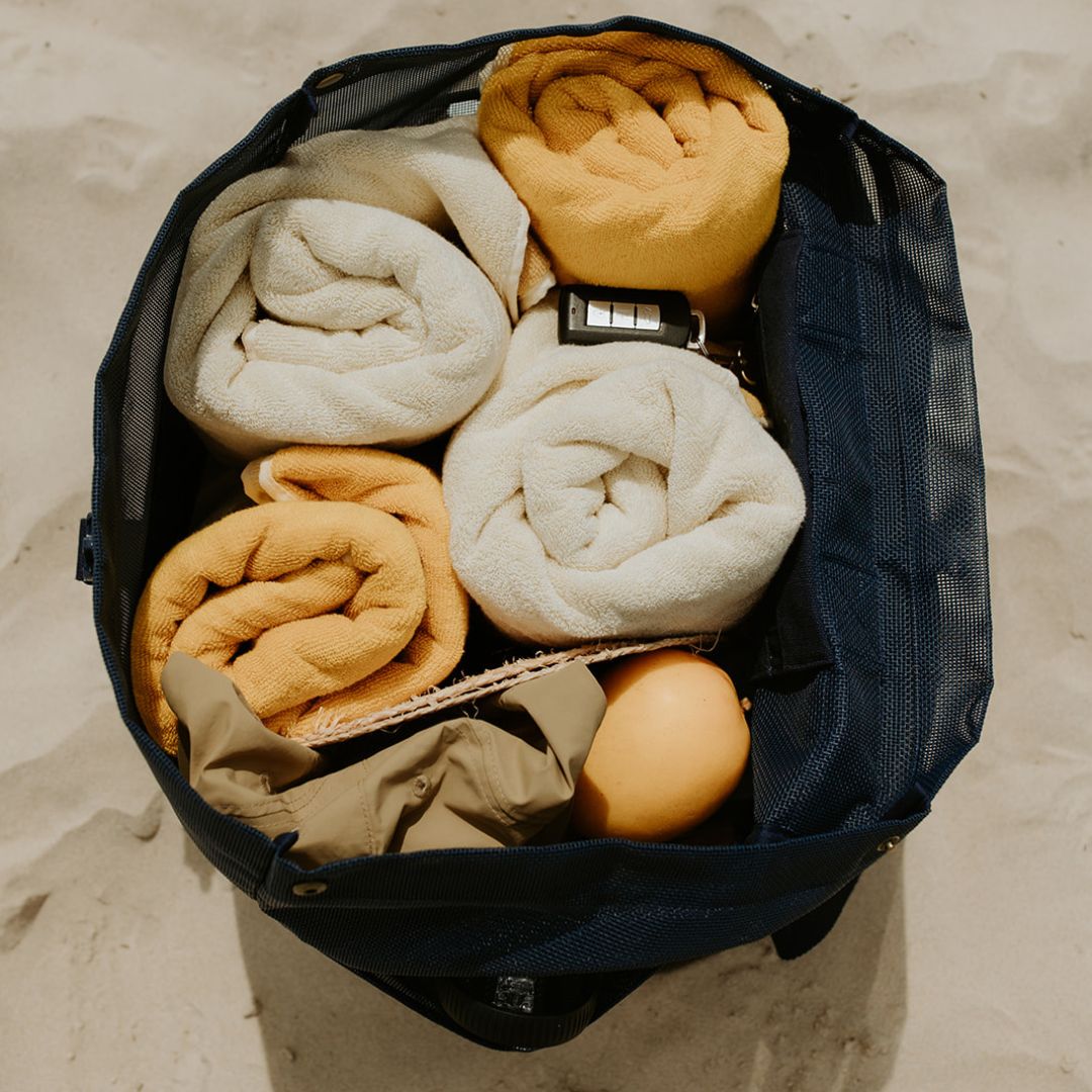 The Sande Beach Hauler mesh backpack in Deep Sea Navy sits on the sand, packed with four bulky beach towels, a hat, sunscreen, snacks, and valuables, demonstrating its large capacity.
