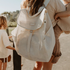 Lifestyle image of mum carrying a toddler on her hip with the Sande  Beach Hauler mesh backpack in Sand on her back, ready for a beach day. Features adjustable straps for comfort.