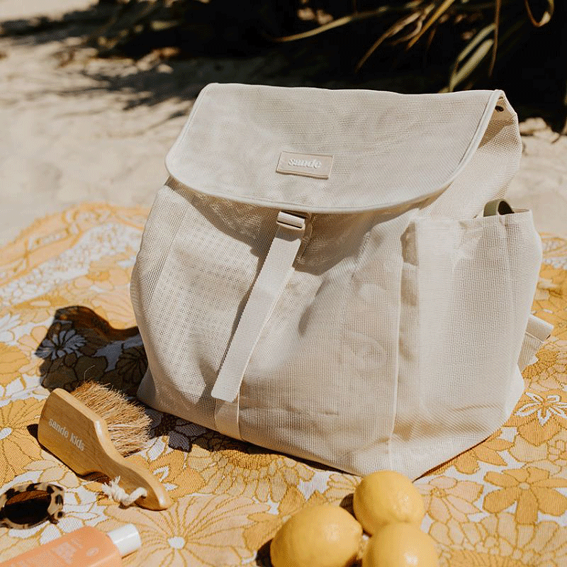 The Sande  Beach Mesh Backpack in Sand, sitting on a yellow picnic blanket next to fresh lemons, sunscreen, sunglasses, and the Sande  Sand Brush. Vegetation is visible in the background.
