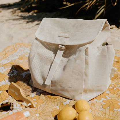 The Sande  Beach Mesh Backpack in Sand, sitting on a yellow picnic blanket next to fresh lemons, sunscreen, sunglasses, and the Sande  Sand Brush. Vegetation is visible in the background.