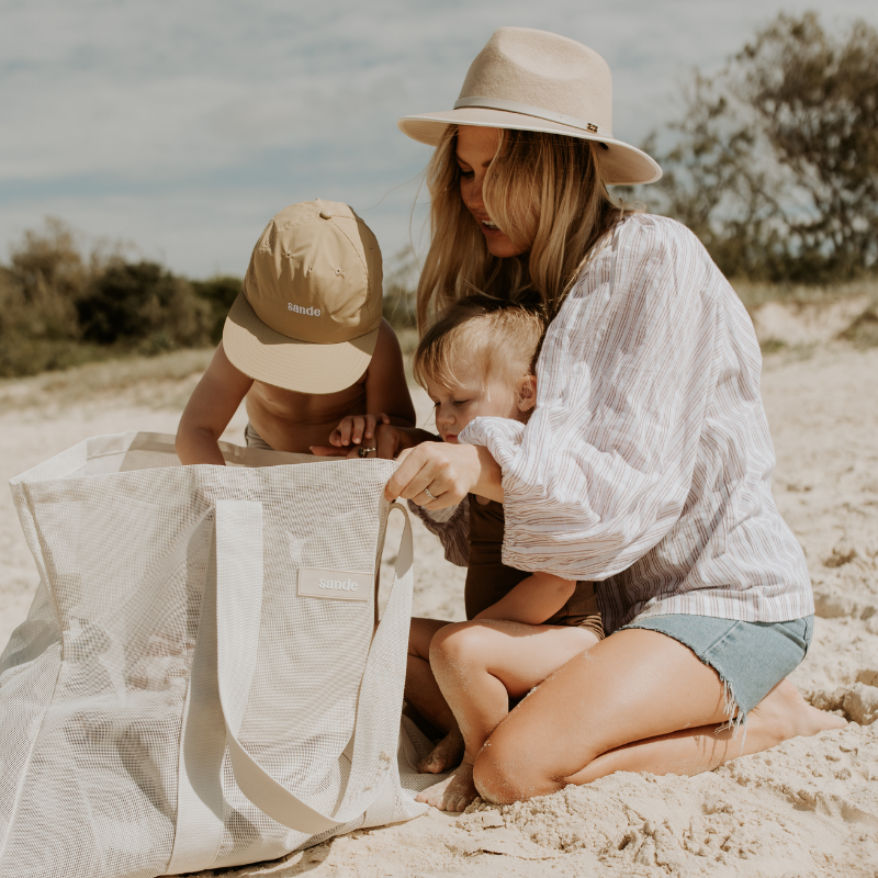 Lifestyle image of a young family looking into their neutral Sande mesh beach bag. A little boy wearing a beige Sande cap reaches into the bag, while a little girl sits on her mothers lap. The mother has long blonde hair and is wearing a white shirt, denim shorts, and a cream fedora hat.