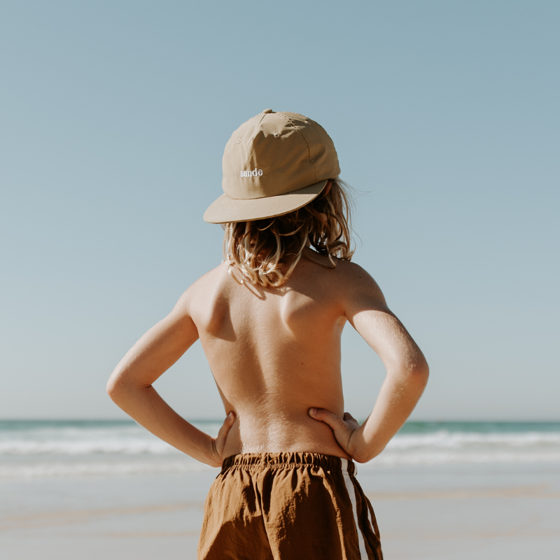 A lifestyle image of a young boy with the beige nylon Sande cap worn backwards. He stands on the beach wearing brown shorts.