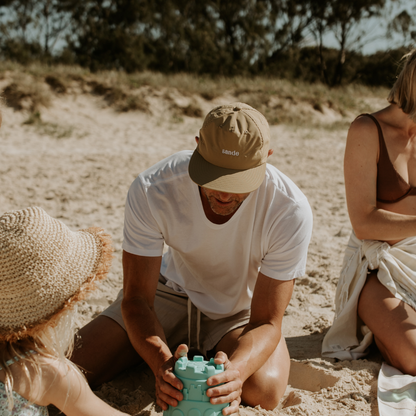 A lifestyle image of dad wearing the beige waterproof Sande cap while building sandcastles on the beach with his family.