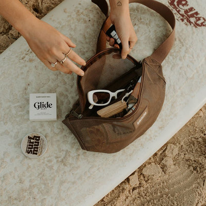 The Sande Essentials Crossbody Bag in Espresso sits on a surfboard as hands reach in. She holds a key in one hand, wearing gold jewellery. Inside the bag are sunglasses, the Sand Brush Mini and other beach essentials including surf wax and surf zinc.