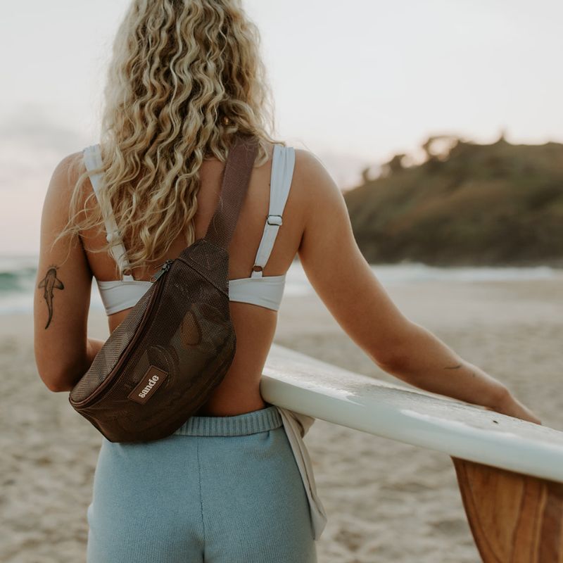 Woman with a Sande™ espresso brown mesh crossbody bumbag and white bikini holding a surfboard on a beach.