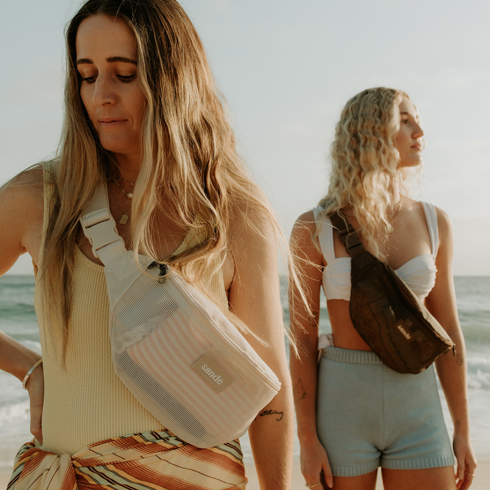 Two women enjoy the beach with the ocean behind them. One wears yellow swimwear with a yellow, red, and green striped sarong and carries the Sande Essentials Crossbody Bag in Sand. Behind her, a woman with long blonde curly hair, tan skin, a white bikini top, and blue shorts wears the Sande Essentials Crossbody Bag in Espresso.
