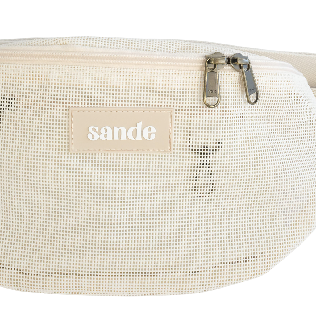 Close up studio detail of the Sande Essentials Crossbody Bag in Sand, highlighting the sand mesh fabric, silicone Sande logo and dual YKK brass zipper pulls on the neutral zip.