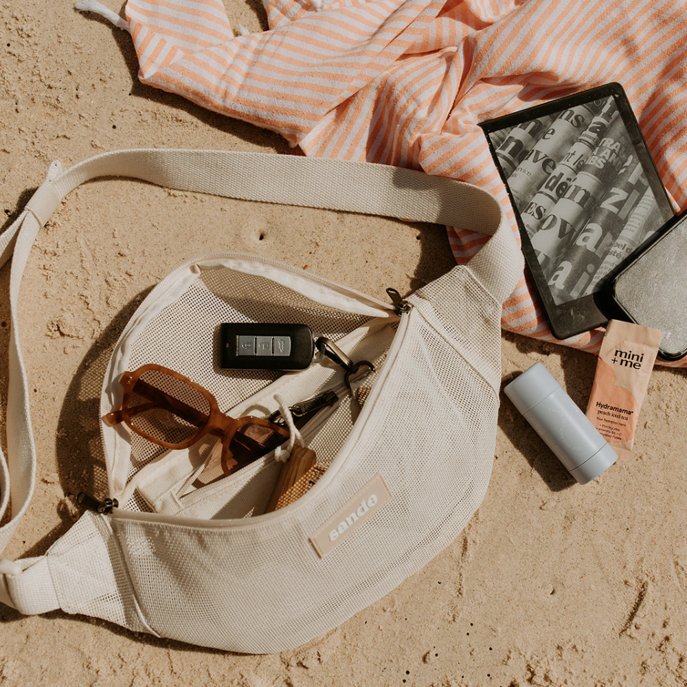 Image of the Sande Essentials Crossbody Bag in Sand, open on the sand next to an orange and white striped Turkish towel. The bag holds sunglasses, keys on the key clip, and the Sand Brush Mini, while a phone, Kindle, and sunscreen rest on the towel.