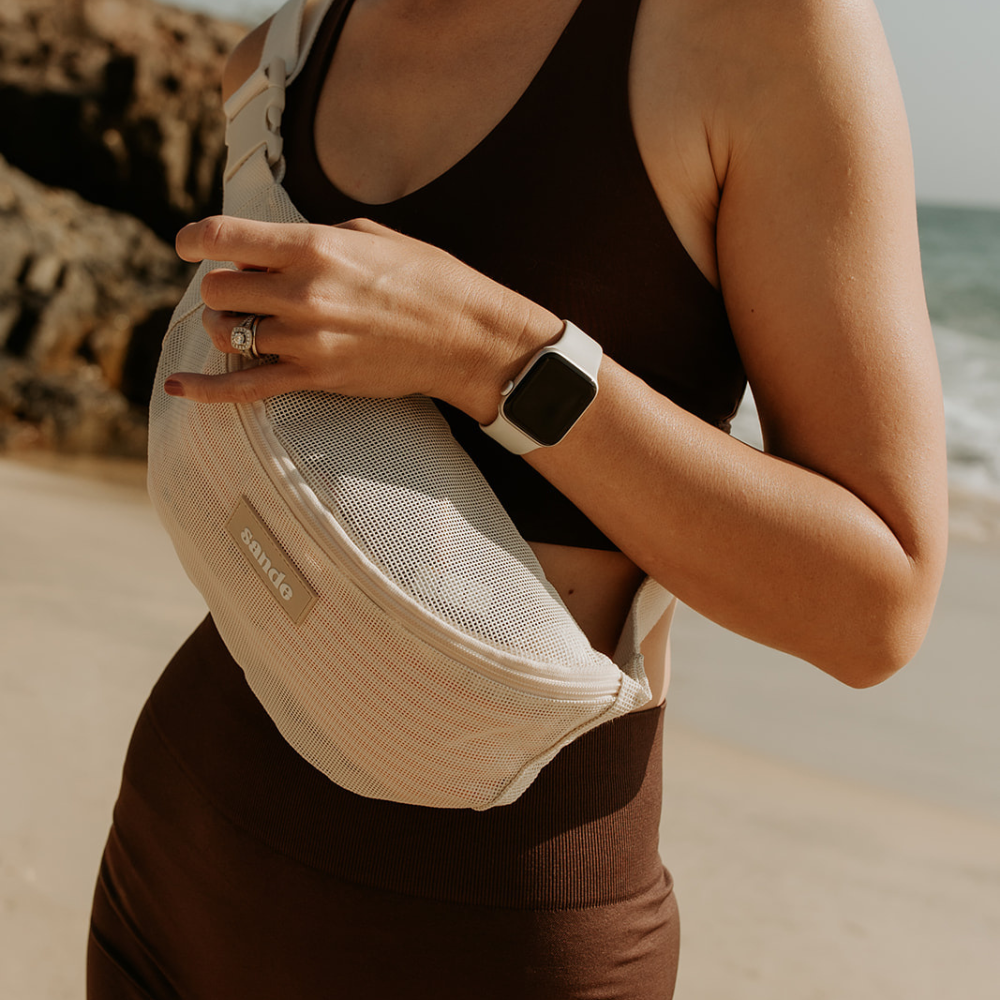 Close-up of a woman at the beach wearing chocolate brown activewear, with the Sande Essentials Crossbody Bag in Sand across her front. Her hand gently holds the top zipper, showing the bag wrapped around her body. The clip on the side and the mesh fabric in the neutral Sand colour are clearly visible.