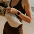 Close-up of a woman at the beach wearing chocolate brown activewear, with the Sande Essentials Crossbody Bag in Sand across her front. Her hand gently holds the top zipper, showing the bag wrapped around her body. The clip on the side and the mesh fabric in the neutral Sand colour are clearly visible.