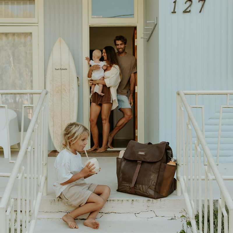 The Sande Beach Hauler Mesh Backpack in Espresso chocolate brown at a beach house, showcasing its stylish design and versatility for beach and coastal lifestyle. The house is pale blue. A mum, dad and baby stand in the doorway, a little boy sipping on a coconut sits on the front step.