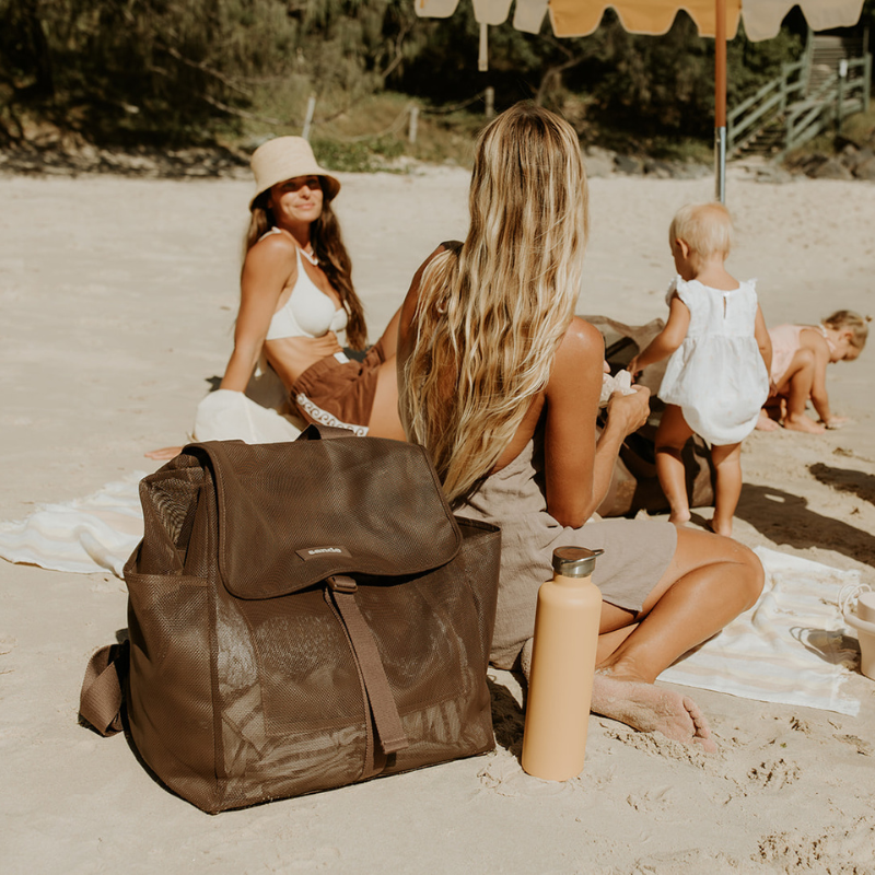 Two women and children on a beach enjoying a beach day with the Sande Beach Hauler Mesh Backpack in Espresso chocolate brown, highlighting its spacious, durable design perfect for group outings and beach gatherings.