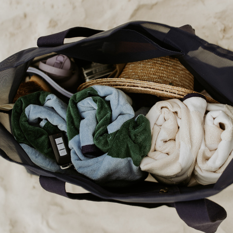 A mum and two little  at the beach, sitting on a green and blue striped beach towel. The mum is rummaging through her oversized mesh beach bag by Sande, the Beach Hauler mesh bag in Deep Sea Navy. The ocean is visible in the background.