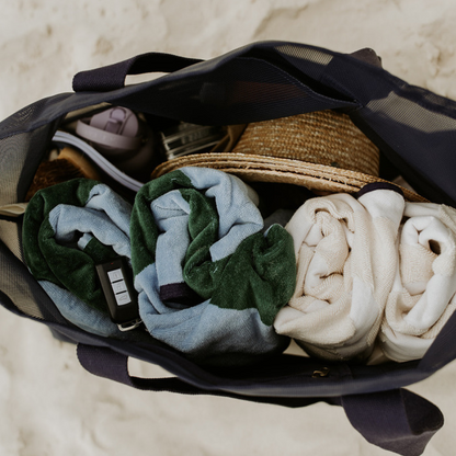 A mum and two little  at the beach, sitting on a green and blue striped beach towel. The mum is rummaging through her oversized mesh beach bag by Sande, the Beach Hauler mesh bag in Deep Sea Navy. The ocean is visible in the background.