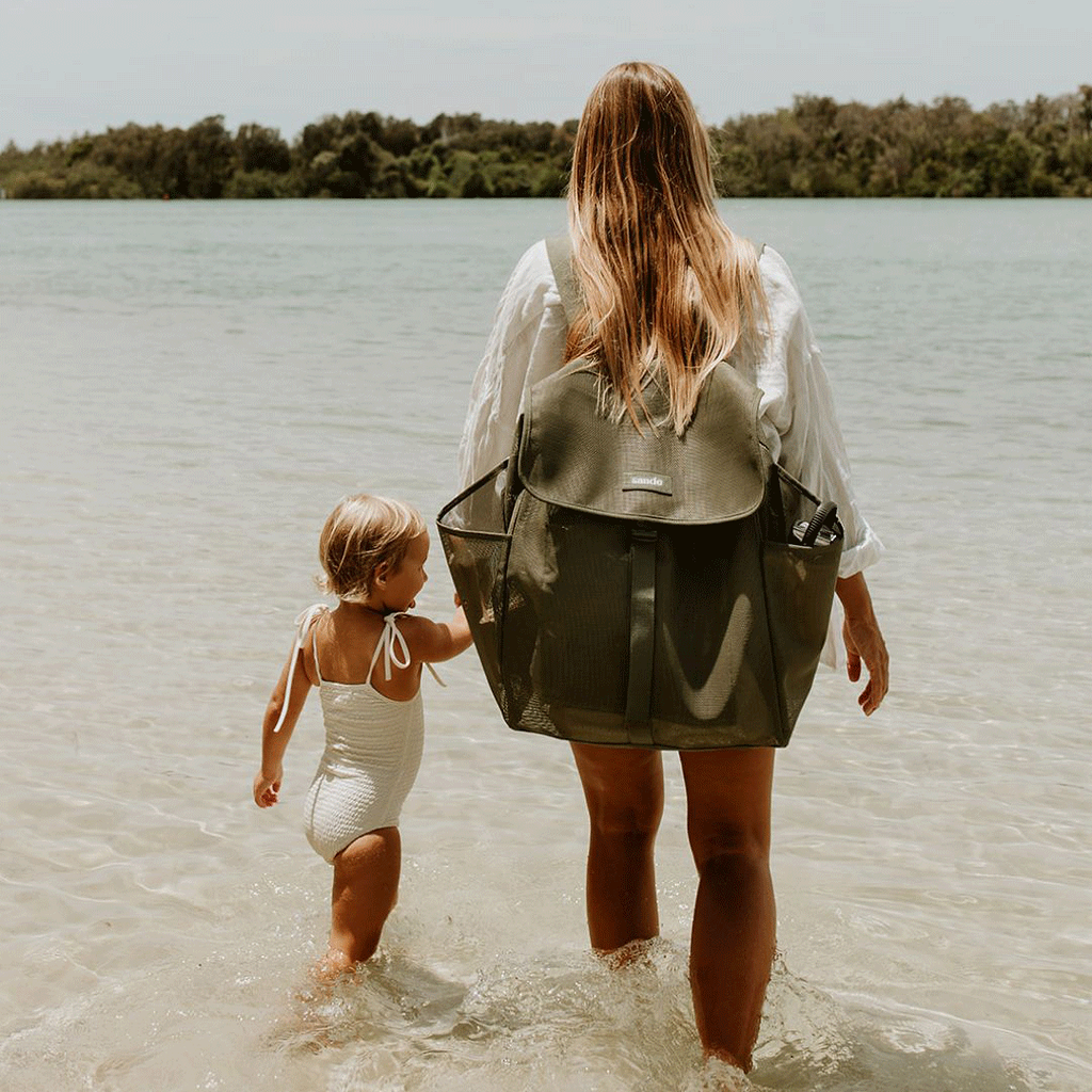 A woman with long blonde hair stands at the waters edge, holding her little girl hand. She wears a dark olive green Sande Beach Hauler mesh backpack, which features side pockets, a flap with a clip, and a white silicone logo. The scene captures a peaceful beach moment, highlighting the backpack style and practicality for family outings.