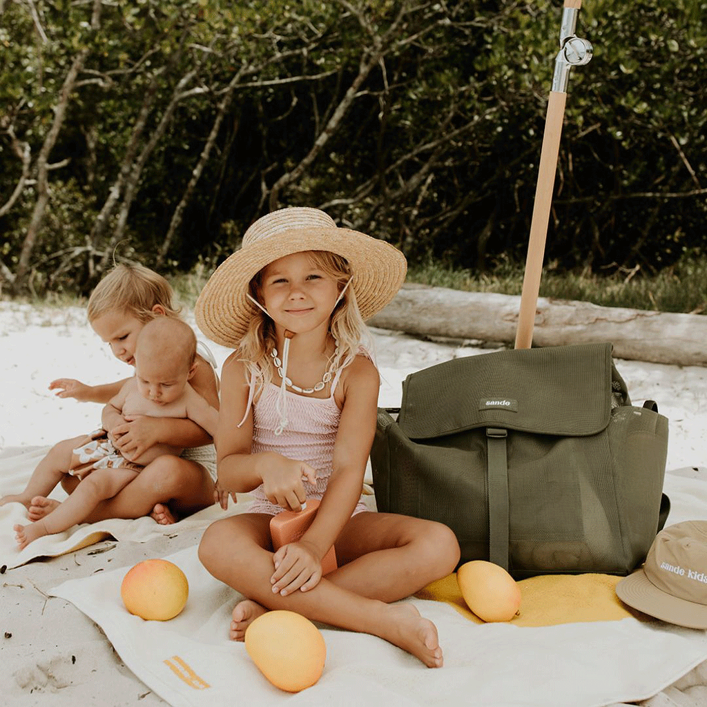 A lifestyle image of young girls sitting under a beach umbrella surrounded by beach towels, sunscreen, and mangoes. One girl wears a wide-brim straw hat and sits next to the Sande Beach Hauler mesh beach backpack in Pandanus Green. The spacious, sand-free backpack features large side mesh pockets for water bottles and toys, adjustable straps, and a durable construction. Perfect for family beach days, outdoor adventures, and carrying essentials while keeping sand out.
