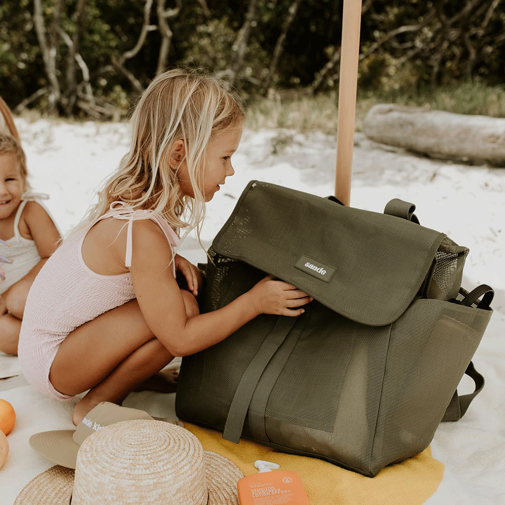 A lifestyle image of a young girl crouching next to the Sande Beach Hauler mesh beach backpack in Pandanus Green. The spacious, sand-free mesh backpack features large side pockets for water bottles and toys, adjustable straps, and a durable construction. Ideal for family beach days, outdoor adventures, and carrying beach essentials while keeping sand out.