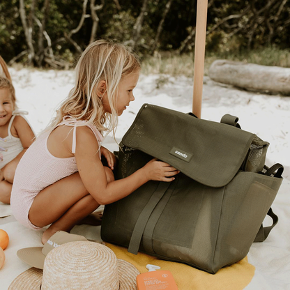 A lifestyle image of a young girl crouching next to the Sande Beach Hauler mesh beach backpack in Pandanus Green. The spacious, sand-free mesh backpack features large side pockets for water bottles and toys, adjustable straps, and a durable construction. Ideal for family beach days, outdoor adventures, and carrying beach essentials while keeping sand out.