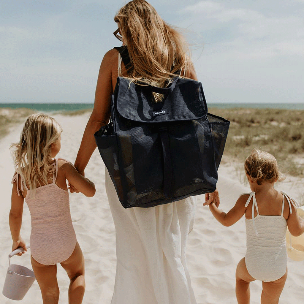 A woman holds the hands of two little girls as they walk along a sandy track to the beach. The mum is wearing the Sande Beach Hauler mesh backpack in Deep Sea Navy.