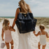 A woman holds the hands of two little girls as they walk along a sandy track to the beach. The mum is wearing the Sande Beach Hauler mesh backpack in Deep Sea Navy.