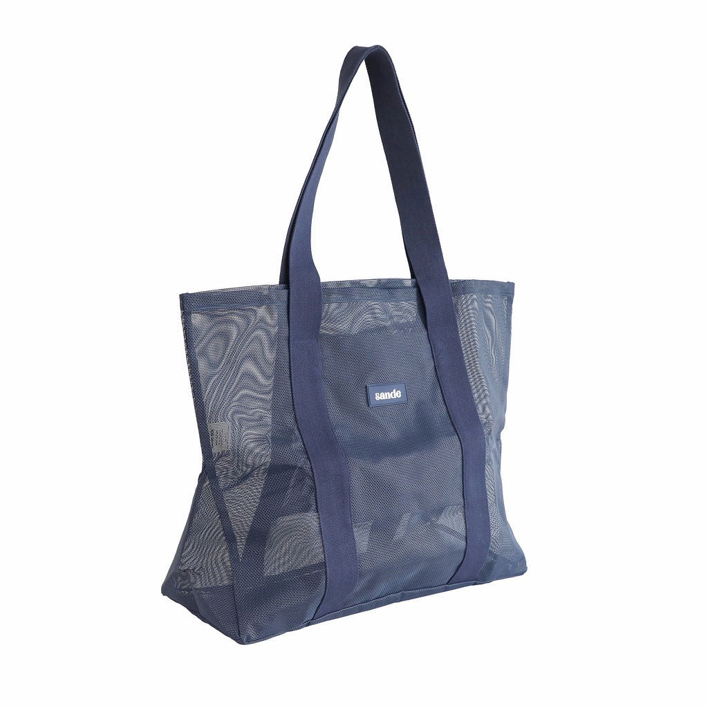 Sande Beach Hauler - large mesh beach bag in Deep Sea Navy, displayed in a studio setting at a 45¬∞ angle. This view highlights the mesh detail, thick cotton straps, durable construction, and the silicone label with the Sande logo. Set against a white background.