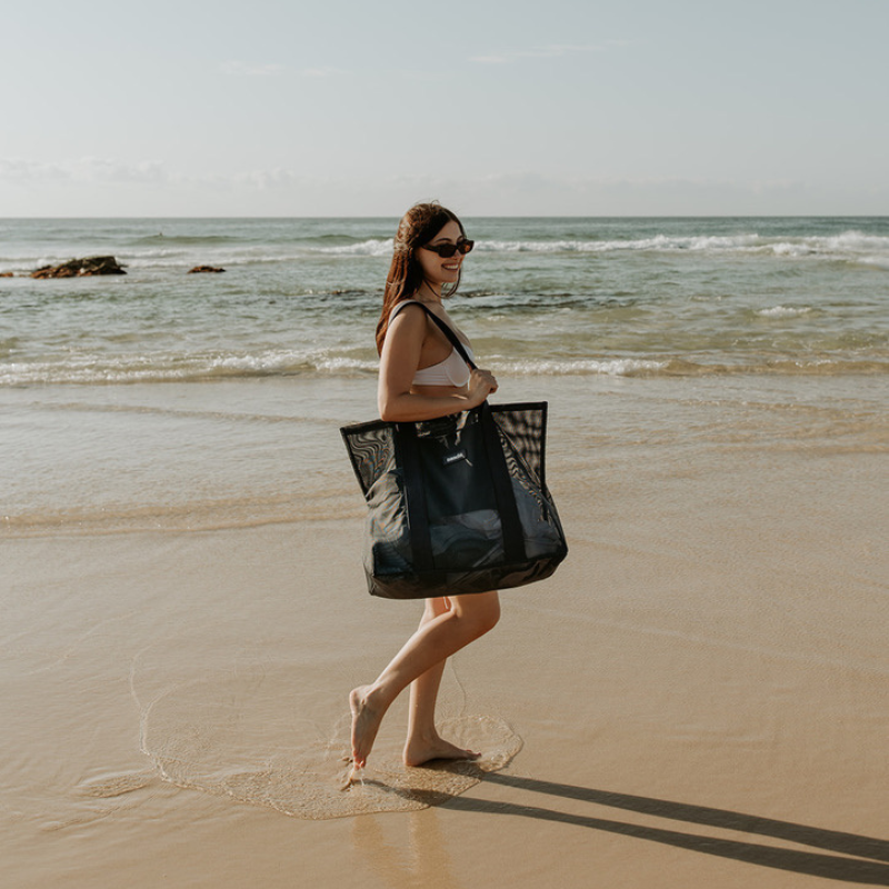 Young woman with long red hair squatting behind the Sande Beach Hauler Mini mesh beach bag in Pandanus Green at the beach, highlighting the spacious size of this large carry-all bag for beach essentials.