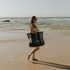 Young woman with long red hair squatting behind the Sande Beach Hauler Mini mesh beach bag in Pandanus Green at the beach, highlighting the spacious size of this large carry-all bag for beach essentials.
