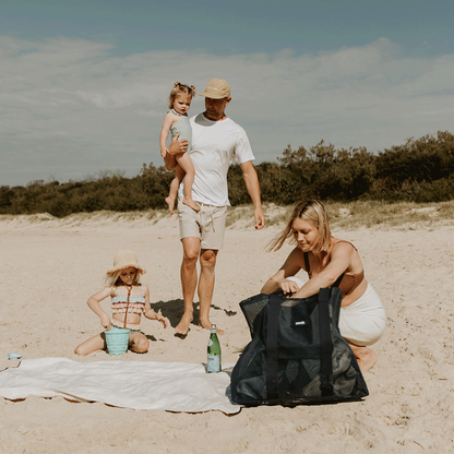 An image of a family at the beach, with a little girl playing with a bucket. Dad is wearing a Sande Cap and carrying a little girl. A beach towel is laid out. The mum, crouching down next to her Deep Sea Navy Beach Hauler mesh beach bag. The beach setting is complemented by grassy dunes and vegetation in the background.