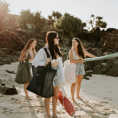 Three friends at the beach, each carrying a Sande Beach Hauler -Mini mesh beach bag in one of our three beautiful colourways: Neutral, Olive Green, and Dark Navy. One girl is carrying a surfboard, while another carries a beach umbrella. They are on the sand with black rocks and pandanus palms  in the background.