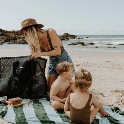 A mum and two little  at the beach, sitting on a green and blue striped beach towel. The mum is rummaging through her oversized mesh beach bag by Sande , the Beach Hauler mesh bag in Deep Sea Navy. The ocean is visible in the background.