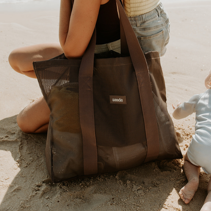 Woman kneeling on the beach beside an espresso brown Sande Kids mesh beach bag tote with Sande branding, with small children nearby.