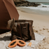 A lifestyle image of the Sande™ Beach Hauler Mini mesh beach bag in Espresso Brown on a sandy beach, accompanied by a straw hat and papayas, with the ocean and rocks in the background. The sand-free, durable mesh bag is perfect for carrying beach essentials for beach days, market trips, and outdoor adventures.
