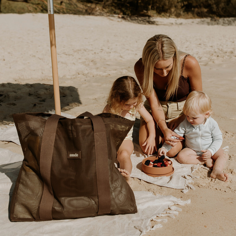 Woman sitting on a sandy beach with her two children beside her, with a brown Sande Beach Hauler mesh beach bag in Espresso.