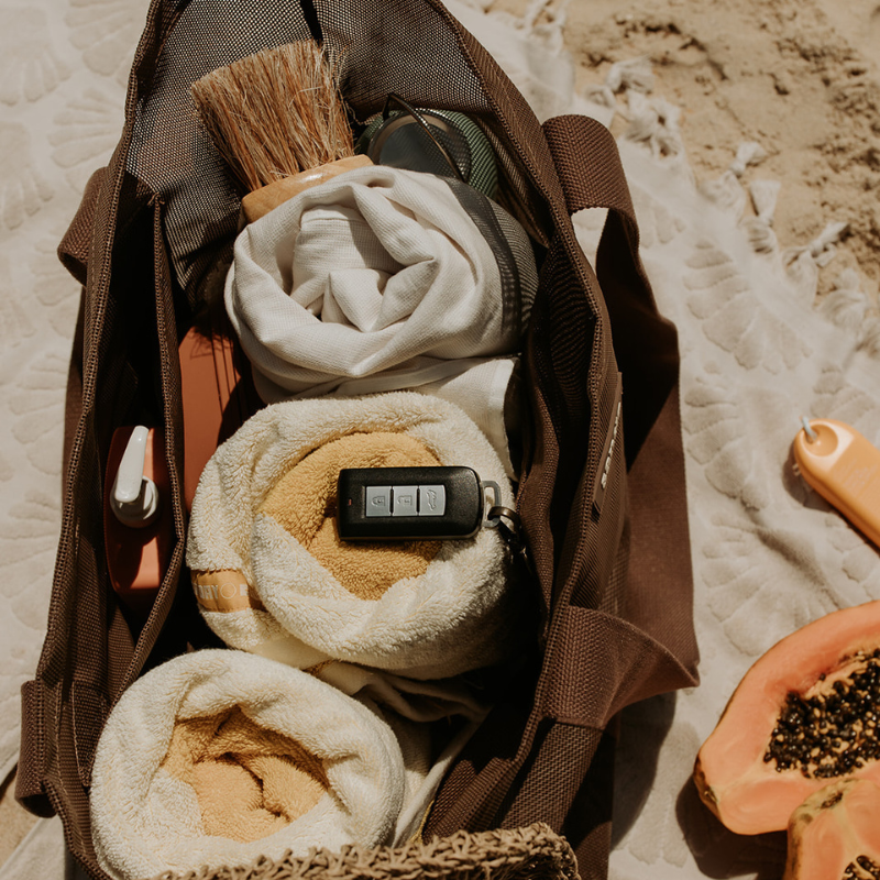 Top view of the Sande mesh beach bag showing capacity with three towels, a sand brush, sunscreen in the internal mesh pocket, and car keys clipped to the key holder. The bag sits on a neutral towel beside fruit and a small yellow sunscreen.