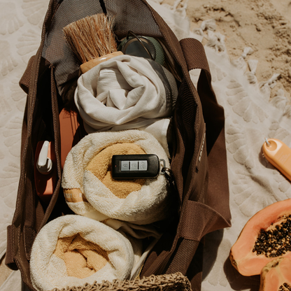 Top view of the Sande mesh beach bag showing capacity with three towels, a sand brush, sunscreen in the internal mesh pocket, and car keys clipped to the key holder. The bag sits on a neutral towel beside fruit and a small yellow sunscreen.