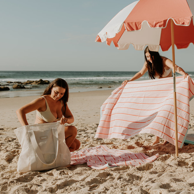 Friends enjoying a day at the beach with their neutral Sande Beach Hauler - Mini beach bags. The scene features pink and red striped beach towels laid out alongside a coral red and white beach umbrella, creating a vibrant and fun atmosphere for a perfect beach day.